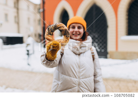Attractive young female tourist in yellow hat is holding pretzel, traditional polish bagel on Market Square in Krakow. Traveling Europe in winter time. High quality photo Attractive young female tourist in yellow hat is holding pretzel, traditional polish bagel on Market Square in Krakow. Traveling Europe in winter time. High quality photo 108705082