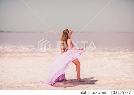 Woman pink salt lake. Against the backdrop of a pink salt lake, a woman in a long pink dress takes a leisurely stroll along the white, salty shore, capturing a wanderlust moment. 108705737