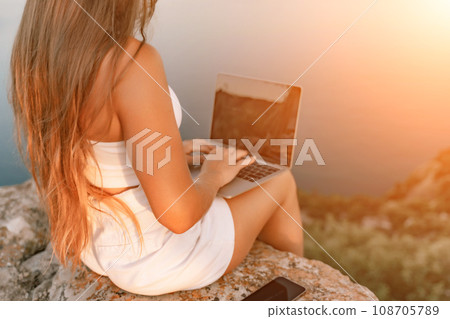 Freelance women sea working on the computer. Good looking middle aged woman typing on a laptop keyboard outdoors with a beautiful sea view. The concept of remote work. 108705789
