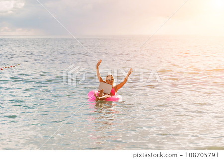 Woman works on laptop in sea. Freelancer, blond woman in sunglases floating on an inflatable big pink donut with a laptop in the sea at sunset. People summer vacation rest lifestyle concept. 108705791