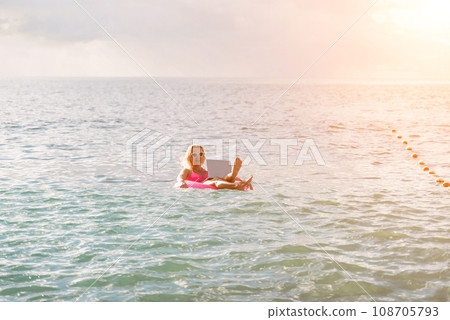 Woman works on laptop in sea. Freelancer, blond woman in sunglases floating on an inflatable big pink donut with a laptop in the sea at sunset. People summer vacation rest lifestyle concept. Woman works on laptop in sea. Freelancer, blond woman in sunglases floating on an inflatable big pink donut with a laptop in the sea at sunset. People summer vacation rest lifestyle concept. 108705793