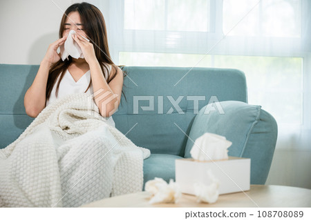 Sick female sitting under blanket on sofa and sneeze with tissue paper in living room, fever caught cold, Asian young woman she sick blowing nose sneezing in tissue at home, medicine healthcare 108708089