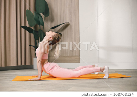 Sporty young woman training at home. Woman wearing rose top and leggins. Woman doing yoga on an orange mat. Sporty young woman training at home. Woman wearing rose top and leggins. Woman doing yoga on an orange mat. 108708124