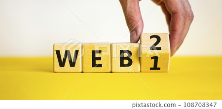 WEB 1 or 2 symbol. Businessman turns a wooden cube and changes words WEB 1 to WEB 2. Beautiful yellow table, white background, copy space. Business, technology and WEB 1 or 2 concept. 108708347