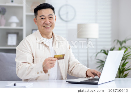 Happy Asian man sitting in bright office at table in front of laptop, holding card in hand, satisfied with online shopping, making online payments. 108710643