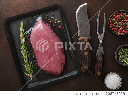 Beef raw fillet steak in vacuum tray with barbeque fork and knife on dark background with spices. Beef raw fillet steak in vacuum tray with barbeque fork and knife on dark background with spices. 108712629