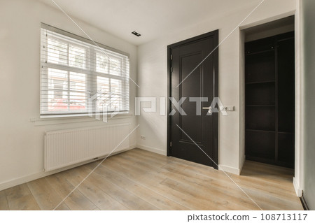 an empty room with white walls and wood flooring, there is a black door that leads to another room an empty room with white walls and wood flooring, there is a black door that leads to another room 108713117