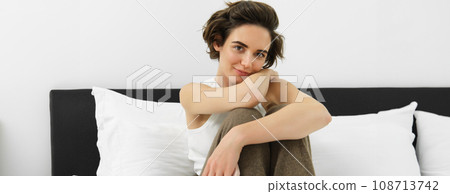 Young tender woman in her cosy pyjamas, sitting on bed in bedroom, smiling with sensual smile Young tender woman in her cosy pyjamas, sitting on bed in bedroom, smiling with sensual smile 108713742