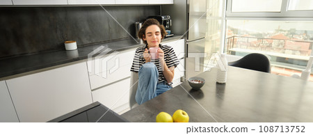 Young woman having breakfast or lunch, eating cereals with milk, drinking hot tea, sitting cosy in the kitchen 108713752