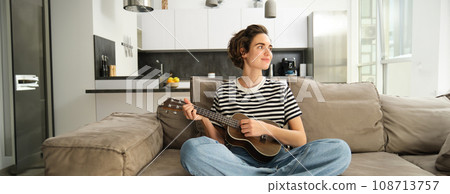 Portrait of girl resting in living room, playing ukulele, learns how to play new musical instrument, spending time relaxing at home Portrait of girl resting in living room, playing ukulele, learns how to play new musical instrument, spending time relaxing at home 108713757