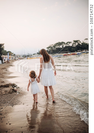 A little girl and a young mother in white dresses walk barefoot on the sand by the sea at sunset.  108714732