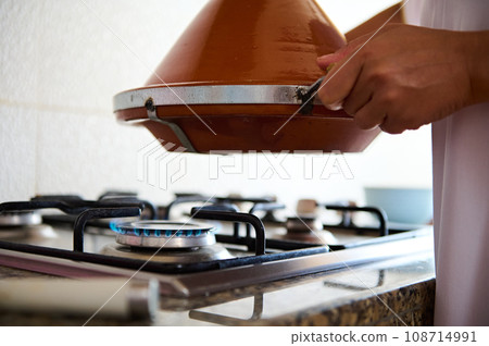 Close-up housewife holding a clay pot tagine above the stove, cooking delicious meal in the home kitchen Close-up housewife holding a clay pot tagine above the stove, cooking delicious meal in the home kitchen 108714991