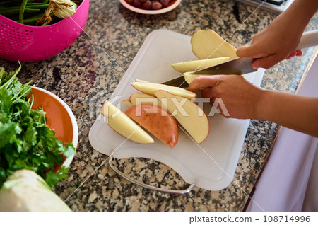Cropped view of female hands slicing potato on a cutting board while preparing dinner in the home kitchen Cropped view of female hands slicing potato on a cutting board while preparing dinner in the home kitchen 108714996