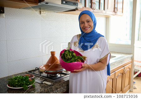 Charming Arabian woman in blue hijab and authentic dress, holds a bowl with washed vegetables, smiles looking at camera 108715016