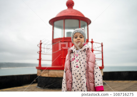 Little girl stands near red lighthouse on rocky seashore 108716275