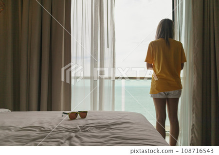 Woman standing at the balcony by the curtain looking out to the sea in vacation. 108716543