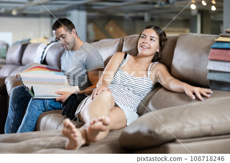 In store wife settled down with legs to test of sofa in use,husband examines upholstery fabric In store wife settled down with legs to test of sofa in use,husband examines upholstery fabric 108718246