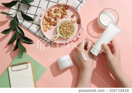 Hand model with a smear on and holding a white tube. Pink dish containing soybeans, almonds and cashew nuts. Unbranded jar displayed. Cosmetic package mock up collection 108718838