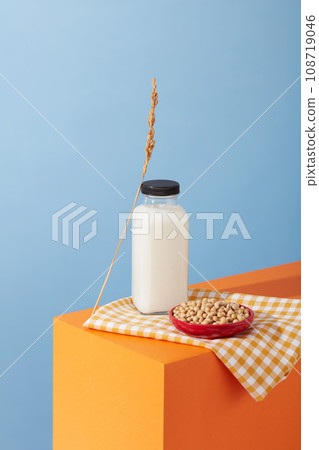 Blank label bottle of milk and a ceramic dish of soybeans are displayed on a white and orange checkered fabric. Nut milk is rich in vitamins and minerals 108719046
