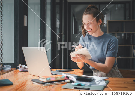 Smiling business woman sitting at office desk and writing notes in a notebook while talking on a video call meeting with clients or coworkers on her laptop computer. Smiling business woman sitting at office desk and writing notes in a notebook while talking on a video call meeting with clients or coworkers on her laptop computer. 108719160
