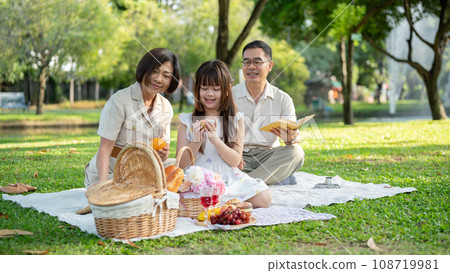 A lovely young Asian girl is enjoying a picnic in the green garden park with her kind grandparents. A lovely young Asian girl is enjoying a picnic in the green garden park with her kind grandparents. 108719981