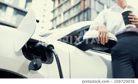 Progressive businesswoman leaning on electric car and charging station. 108720036