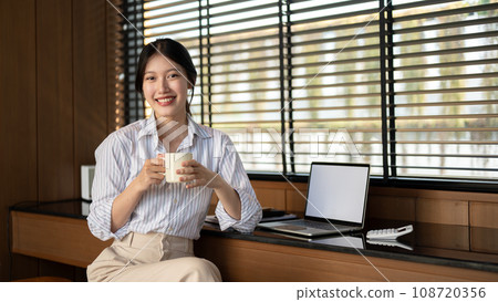 A beautiful Asian businesswoman is sitting at a table indoors with a coffee mug in her hands. 108720356