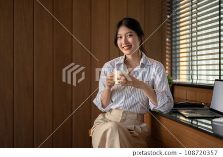 A beautiful Asian businesswoman is sitting at a table indoors with a coffee mug in her hands. 108720357
