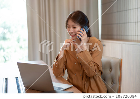 A charming Asian businesswoman is talking on the phone with her client while working in her office. 108720384