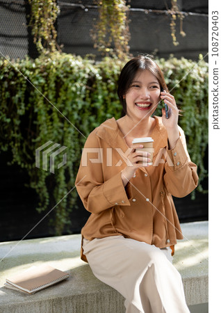 A happy Asian woman is enjoying talking on the phone and sipping coffee while relaxing on a bench. A happy Asian woman is enjoying talking on the phone and sipping coffee while relaxing on a bench. 108720403
