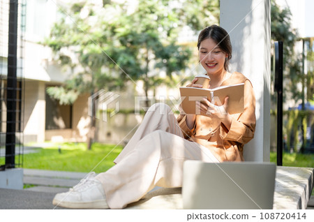 A beautiful Asian woman is sipping coffee and enjoying reading a book on a bench in a garden. 108720414