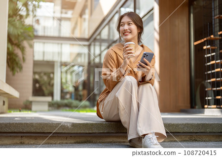 Beautiful Asian woman is sitting on the stairs with her phone and a takeaway coffee cup in her hands Beautiful Asian woman is sitting on the stairs with her phone and a takeaway coffee cup in her hands 108720415