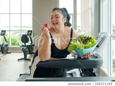 A plus-sized lady is exercising on a treadmill, maintaining her health by having a bowl of fresh fruit and vegetable. 108721184