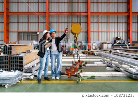 Two women are standing inside a big warehouse, examining a tall crane attentively. Two women are standing inside a big warehouse, examining a tall crane attentively. 108721233