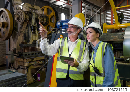 Senior engineer manager trains new employees within the metal sheet factory. Everyone wear safety vest and hardhat. Various kind of industrial machine are in the working area. 108721515