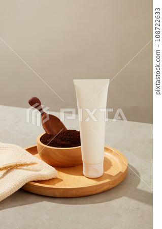 Front view of plastic tube unlabeled on round wooden dishes with coffee powder on wooden bowl and towel on gray background. Mockup for cosmetic, facial moisturizer cream or facial cleanser. 108722633