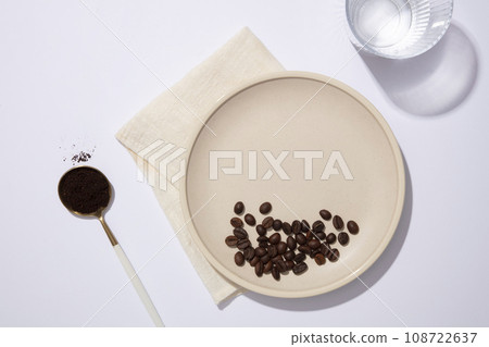 Against the white background, coffee beans and coffee powder contained on ceramic dish and spoon, decorated with glass cup and beige fabric. Blank space on dish for product presentation 108722637