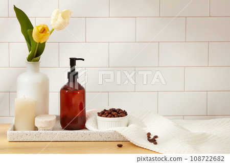 An amber pump bottle displayed with bowl of coffee beans, candle and vase of flower on marble tray on white ceramic tile background. Space for design packaging on bottle. Advertising photo 108722682