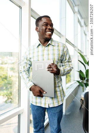Young african man standing near the window indoors and holding laptop Young african man standing near the window indoors and holding laptop 108726207