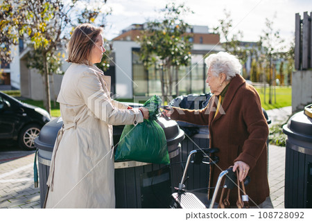 Woman helping elderly neighbor throw away trash into garbage can, waste container in front of their apartment complex. 108728092
