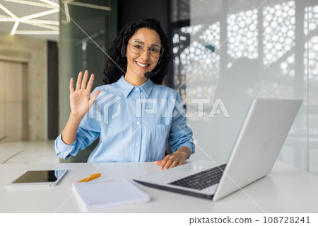 Friendly happy hispanic businesswoman in headset working in call center sitting at desk in front of laptop, young brunette office worker making video call, waving hello to camera. Friendly happy hispanic businesswoman in headset working in call center sitting at desk in front of laptop, young brunette office worker making video call, waving hello to camera. 108728241