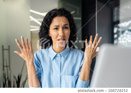 Dissatisfied office worker Indian woman sitting in the office in front of the laptop spreads her hands in confusion, problems with work, non-working laptop, stressful situation at work. Dissatisfied office worker Indian woman sitting in the office in front of the laptop spreads her hands in confusion, problems with work, non-working laptop, stressful situation at work. 108728252