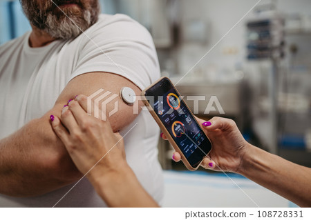 Doctor connecting continuous glucose monitor with smartphone, to check blood sugar level in real time. Obese, overweight man is at risk of developing type 2 diabetes. Concept of health risks of 108728331