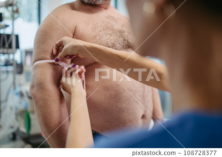 Female doctor measuring arm circumference of overweight patient using tape measure. Obesity affecting middle-aged men's health. Concept of health risks of overwight and obesity. 108728377