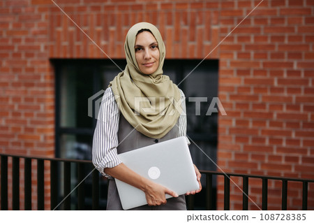 Beautiful woman in hijab standing on city street. Muslim businesswoman with laptop in hands. Iran, Afganistan female teacher in front of school building. Beautiful woman in hijab standing on city street. Muslim businesswoman with laptop in hands. Iran, Afganistan female teacher in front of school building. 108728385