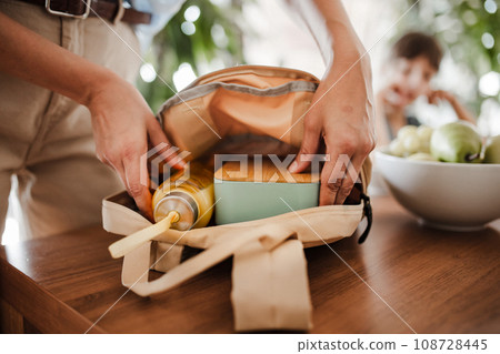 Mother packing school bag for kid. Putting bottle of juice, snacks for children into school bag. Snack, school lunch in a snack box for school. Mother packing school bag for kid. Putting bottle of juice, snacks for children into school bag. Snack, school lunch in a snack box for school. 108728445