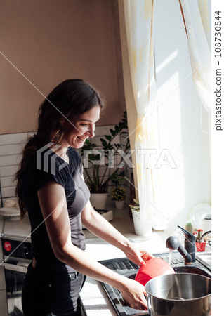 a woman washes dishes in the kitchen by the window a woman washes dishes in the kitchen by the window 108730844