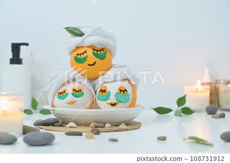 Orange fruits in face mask, zen stones, candles and green leaves on white background. Spa treatment and self care concept Orange fruits in face mask, zen stones, candles and green leaves on white background. Spa treatment and self care concept 108731912