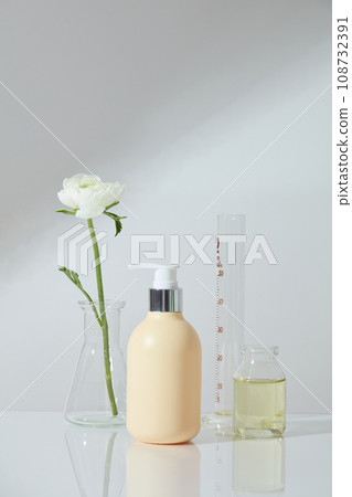 A jar of essential oil, measuring cylinder and a conical flask with flower displayed with an unlabeled pump bottle. Front view. Cosmetic laboratory research and development A jar of essential oil, measuring cylinder and a conical flask with flower displayed with an unlabeled pump bottle. Front view. Cosmetic laboratory research and development 108732391
