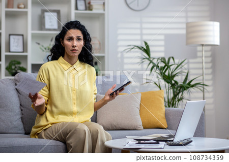 Curly upset Indian woman businesswoman working from home, sitting in front of laptop on sofa doing paper work, tired of many tasks, burnout, overwork. Curly upset Indian woman businesswoman working from home, sitting in front of laptop on sofa doing paper work, tired of many tasks, burnout, overwork. 108734359